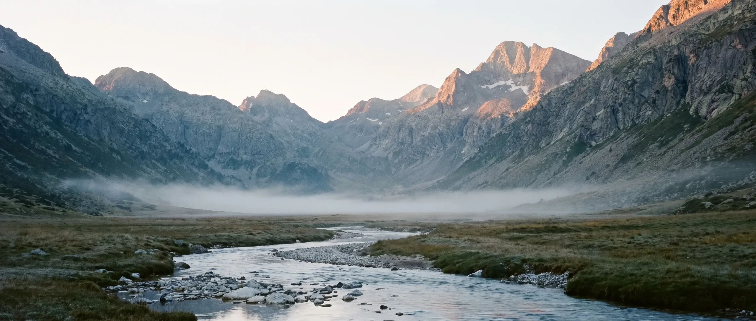 A high Pyrenean valley at first light, mist lying low and a meltwater stream running through the foreground